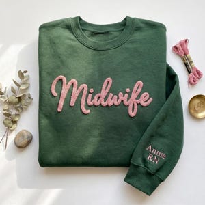 Op de afbeelding: Een donkergroene sweater met ronde hals en het woord Midwife in verhoogde, textuurrijke roze schrijfletters op de borst. De rechtermouwboord is geborduurd met de naam Annie en de letters RN in bijpassend roze garen.