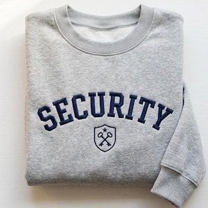 Könnte beinhalten: Ein gefalteter hellgrauer Sweatshirt mit dem Wort "SECURITY" in Marineblau über der Brust gestickt. Darunter befindet sich ein marineblaues Schild mit gekreuzten Schlüsseln und einem Stern.