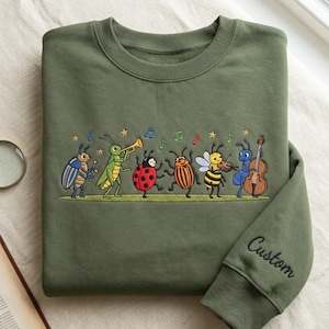 Könnte beinhalten: Ein olivgrünes Sweatshirt mit Rundhalsausschnitt und einer gestickten Szene von sechs bunten Insekten, die Musikinstrumente wie Trompete, Geige und Cello spielen, begleitet von Noten und Sternen. Das Wort Custom ist auf dem Ärmel eingestickt.