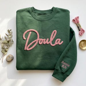 Może przedstawiać: Ciemnozielona bluza z napisem "Doula" wyhaftowanym różową czcionką na piersi. Na rękawie widnieje napis "Annie RN" w kolorze różowym. Bluza leży płasko na białej powierzchni, w pobliżu różowa nić i małe złote pierścienie.