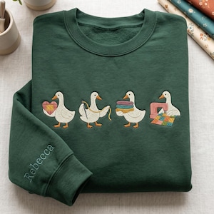 May include: A dark green sweatshirt with embroidered geese, each holding a craft item: a heart, needle and thread, fabric, and a sewing machine. The name "Rebecca" is embroidered on the sleeve.