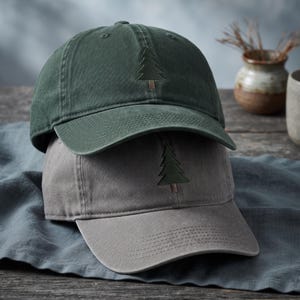 May include: A dark green baseball cap stacked on top of a grey baseball cap. Both caps feature a simple embroidered evergreen tree design on the front centre panel. The caps have a structured crown, a curved brim, and visible stitching details.
