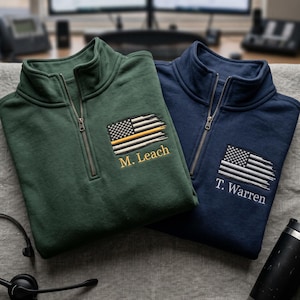 Könnte beinhalten: Ein dunkelgrüner Pullover mit kurzem Reißverschluss und einer gestickten, abgenutzten amerikanischen Flagge mit einer dünnen gelben Linie und dem Namen M. Leach, neben einem marineblauen Pullover mit weißer Flagge und dem Namen T. Warren.