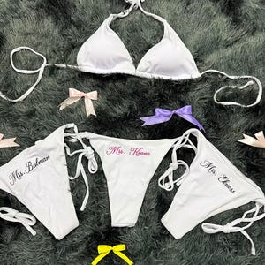 May include: White bikini set with a triangle top and three thong bottoms, each embroidered with a different name in pink or black. The set includes several small bows in various colors.
