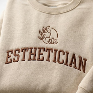 May include: Beige crewneck sweatshirt with the word "ESTHETICIAN" embroidered in brown, arched across the chest. Above the text is a brown embroidered design of a woman's profile, leaves, and a lotus flower.