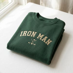 May include: A folded dark green sweatshirt featuring the embroidered text "IRON MAN" in a beige, arched font. Below the text is a graphic of crossed golf clubs and a golf ball. The sweatshirt is neatly folded on a white surface.