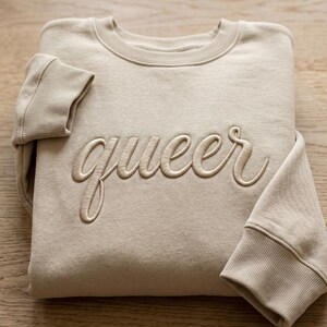 May include: A beige sweatshirt with the word "queer" embroidered in a matching colour. The sweatshirt is folded, with the sleeves visible. The fabric appears soft and comfortable. The design is simple and elegant.