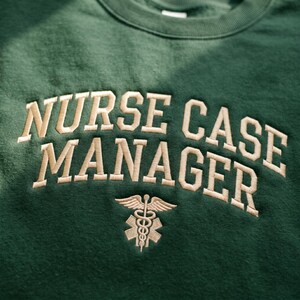 Könnte beinhalten: Ein dunkelgrünes Sweatshirt mit den Worten "NURSE CASE MANAGER" in hellbrauner Farbe gestickt. Unter dem Text befindet sich ein medizinisches Symbol mit einem Caduceus und einem Stern.