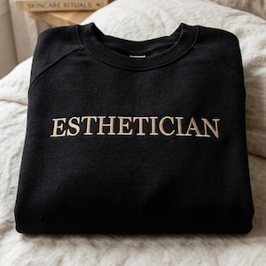 May include: Black sweatshirt with the word "ESTHETICIAN" embroidered in a light tan color across the chest. The sweatshirt is folded on a light-colored surface.