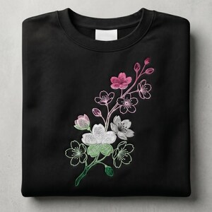 May include: Black sweatshirt featuring an embroidered floral design. The design includes pink, white, and green blossoms with delicate outlines. The sweatshirt has a crew neck and a blank label.