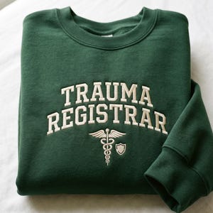 May include: A dark green sweatshirt with the words "TRAUMA REGISTRAR" embroidered in a cream-colored, collegiate-style font. Below the text is a cream-colored medical symbol and shield.
