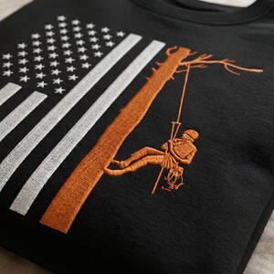 May include: Black sweatshirt featuring an embroidered design. The design includes an American flag with white stars and stripes, and an orange tree with a person climbing it. The person is wearing safety gear.