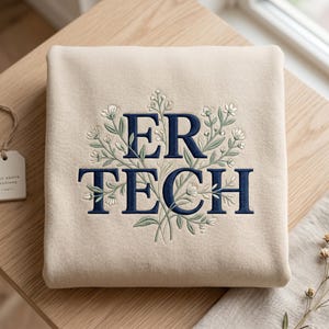 May include: A beige sweatshirt featuring dark blue embroidered text that reads ER TECH. The lettering is surrounded by detailed embroidery of green stems and small white flowers, creating a botanical design centered on the front of the garment.