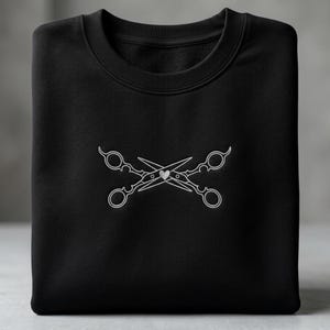 Op de afbeelding: Zwart sweatshirt met een ronde hals. De voorkant heeft een wit geborduurd ontwerp van gekruiste scharen met een klein hartje in het midden. De scharen hebben sierlijke handvatten. Het sweatshirt is netjes opgevouwen.