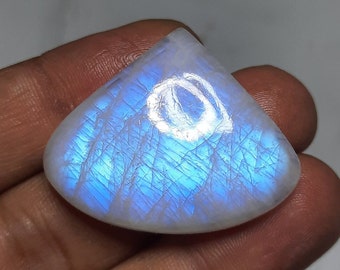 May include: A polished, triangular moonstone gemstone. The stone has a pearly white base with internal blue and white iridescent patterns. The surface reflects light, creating a soft glow. The gemstone is held in a hand.