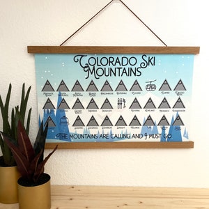 May include: A blue and white poster with a list of Colorado ski mountains. The poster has a wooden frame and is hanging on a white wall. The text on the poster reads "Colorado Ski Mountains" and "The Mountains Are Calling And I Must Go."