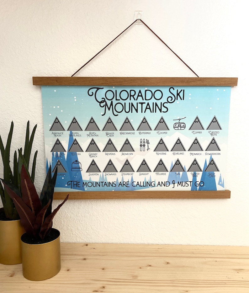 May include: A blue and white poster with a list of Colorado ski mountains. The poster has a wooden frame and is hanging on a white wall. The text on the poster reads "Colorado Ski Mountains" and "The Mountains Are Calling And I Must Go."
