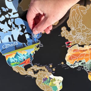 May include: A black world map with gold foil detailing, featuring country outlines and names. A clear, circular tool is being used to scratch off a section of the map, revealing a colorful design beneath. The map includes the text "Canada" and "U.S.A."