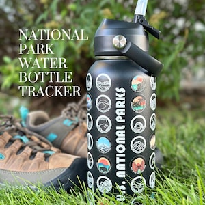 May include: Black water bottle with a black lid and a black handle. The bottle has a sticker on it that says "US National Parks" and has a design of different national parks. The bottle is sitting on green grass.