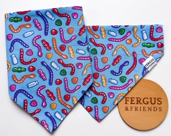 May include: Two blue dog bandanas with a pattern of colourful gummy sweets. The bandanas are triangular and feature gummy worms and other candy shapes in red, orange, purple, and green. The brand name "FERGUS & FRIENDS" is visible.
