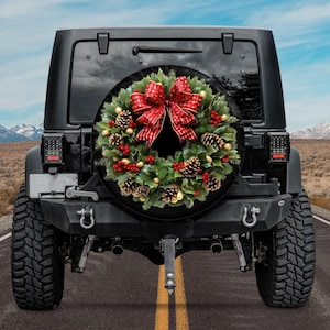 Puede incluir: Un Jeep negro con una cubierta de rueda de repuesto que presenta una corona de Navidad con un lazo rojo, piñas y bayas de acebo.