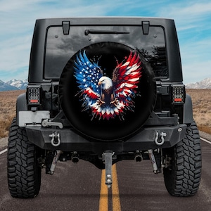 Puede incluir: Un Jeep negro con una cubierta de rueda de repuesto que presenta un águila calva americana con las alas extendidas, sosteniendo la bandera estadounidense en sus garras. El águila está rodeada de salpicaduras de pintura roja, blanca y azul.
