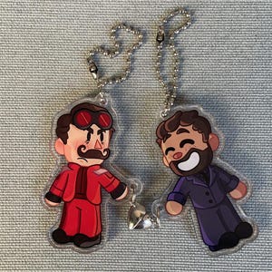 Pode incluir: Dois chaveiros de personagens de desenhos animados com correntes prateadas. O chaveiro à esquerda apresenta um personagem com um fato vermelho, bigode e óculos vermelhos. O chaveiro à direita apresenta um personagem com um fato roxo, barba e um grande sorriso.