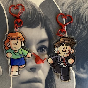May include: Two cartoon character keychains with red heart-shaped clasps. One keychain features a character in a green shirt and blue skirt. The other keychain depicts a character in a suit with blood splatter. Both have a distressed look.