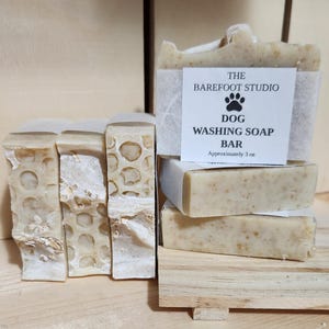 Puede incluir: Barras de jabón para lavar perros de The Barefoot Studio. Las barras son de color beige claro con avena visible y pesan aproximadamente 85 g. Varias barras están apiladas sobre una pequeña plataforma de madera, otras de pie.