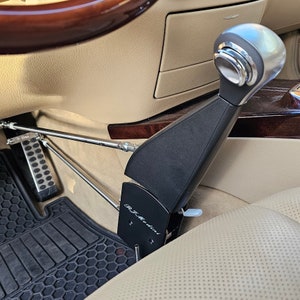 May include: A black and silver car hand control system, with the text "R.J. Modini" on the side. The system is installed in a car with tan leather seats and a black floor mat. The gear shift is silver and black.
