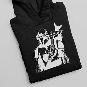Puede incluir: Sudadera con capucha negra con un gr&aacute;fico blanco de un personaje mandaloriano llevando una peque&ntilde;a criatura similar a un ni&ntilde;o en su espalda.