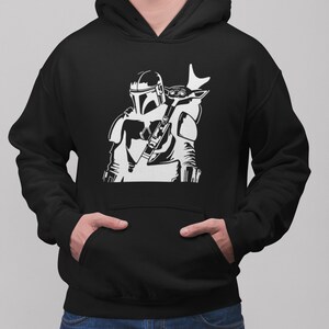 Puede incluir: Sudadera con capucha negra con un estampado gr&aacute;fico blanco de un personaje mandaloriano de Star Wars que lleva una peque&ntilde;a criatura similar a un ni&ntilde;o en la espalda.