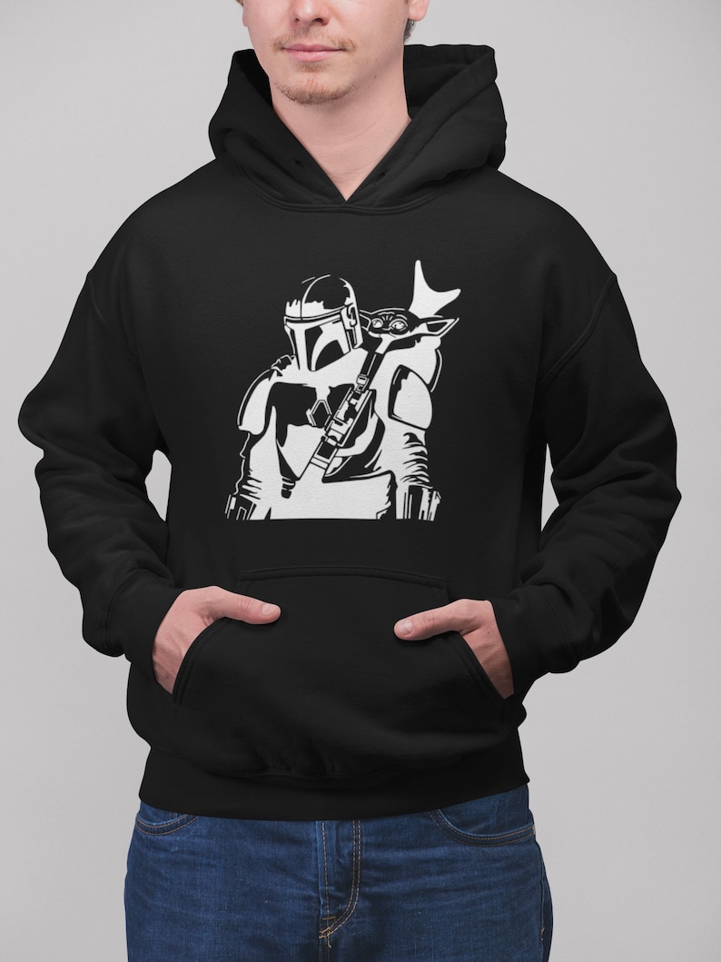 Puede incluir: Sudadera con capucha negra con un estampado gr&aacute;fico blanco de un personaje mandaloriano de Star Wars que lleva una peque&ntilde;a criatura similar a un ni&ntilde;o en la espalda.