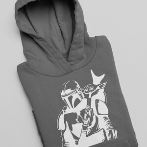 Puede incluir: Una sudadera con capucha gris con una silueta blanca de un personaje mandaloriano que lleva una peque&ntilde;a figura infantil en su hombro.