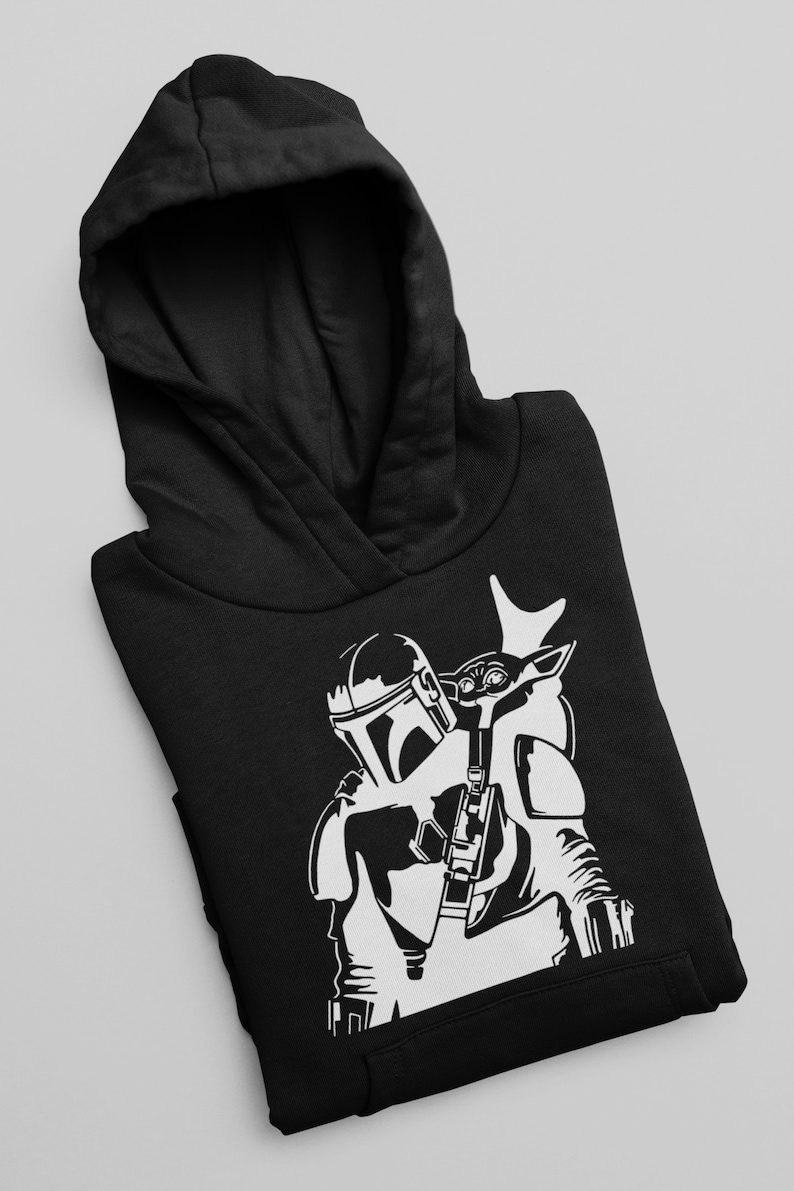 Puede incluir: Sudadera con capucha negra con un gr&aacute;fico blanco de un personaje mandaloriano llevando una peque&ntilde;a criatura similar a un ni&ntilde;o en su espalda.