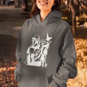 Puede incluir: Una sudadera con capucha gris con una silueta blanca de un personaje mandaloriano llevando una peque&ntilde;a criatura en la espalda.