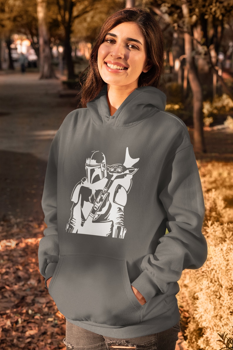 Puede incluir: Una sudadera con capucha gris con una silueta blanca de un personaje mandaloriano llevando una peque&ntilde;a criatura en la espalda.
