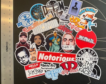 Può includere: Una collezione di vari adesivi in vinile a tema hip-hop con artisti, loghi e testi come Notorious, Slim Shady e Ruthless Records, disposti accanto a un righello metallico che misura fino a 18 cm di lunghezza su una superficie scura.