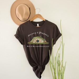 May include: A brown t-shirt with a white graphic of a hobbit hole with the text "Merry & Pippin's Bed & Second Breakfast".