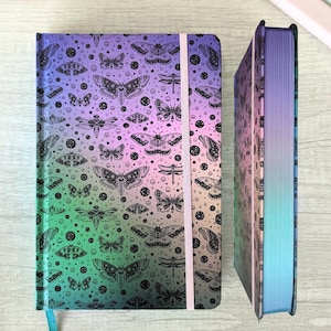 May include: Two hardback journals with a gradient cover in shades of green, purple, and blue. The covers feature a black moth and dragonfly pattern. One journal is open, revealing the edge of the pages and a blue ribbon bookmark.