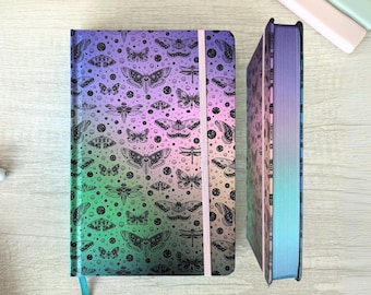 May include: Two hardback journals with a gradient cover in shades of green, purple, and blue. The covers feature a black moth and dragonfly pattern. One journal is open, revealing the edge of the pages and a blue ribbon bookmark.