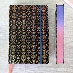 May include: Two notebooks, one with a black cover and a gold skull pattern, and the other with a pink to purple gradient cover. Both notebooks have elastic bands and ribbon bookmarks.