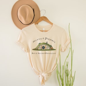 May include: A beige t-shirt with a graphic of a hobbit hole with the text "Merry & Pippin's Bed & Second Breakfast" on it.