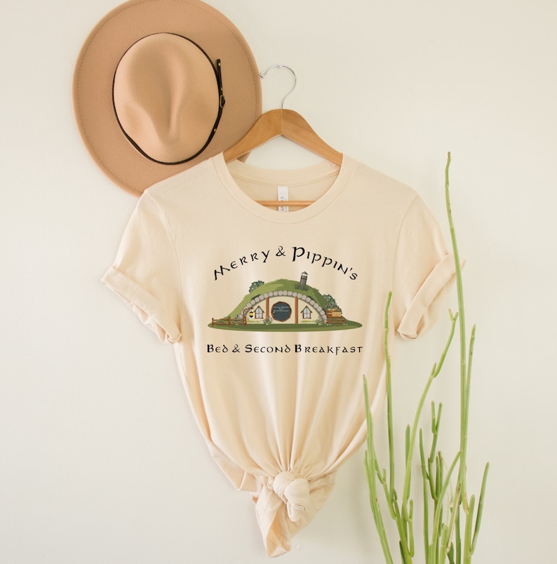 May include: A beige t-shirt with a graphic of a hobbit hole with the text "Merry & Pippin's Bed & Second Breakfast" on it.