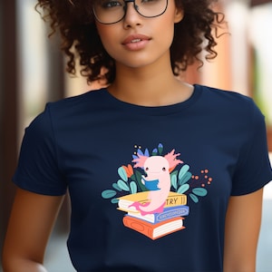 May include: A navy blue t-shirt with a pink axolotl reading a book on top of a stack of books. The books are labeled "TRY II" and "ENCYCLOPEDIA".