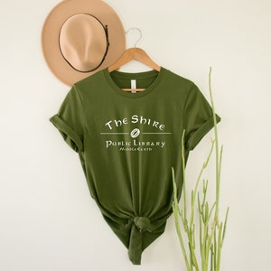Op de afbeelding: Een groen T-shirt met witte tekst die "The Shire Public Library Middle-earth" luidt. De tekst is in een gestileerd lettertype en bevat een klein beeld van een ring.