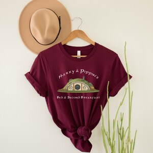May include: A maroon t-shirt with a white graphic of a hobbit hole with the text "Merry & Pippin's Bed & Second Breakfast".
