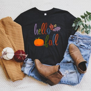 Puede incluir: Camiseta negra con el texto "hello fall" en letras de colores, una pequeña calabaza naranja y hojas de colores. La camiseta está sobre una superficie blanca con un suéter marrón, jeans azules y botas marrones.