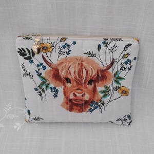 May include: A white zippered pouch featuring a watercolor illustration of a Highland cow with a floral design. The cow is brown with long hair, surrounded by yellow and blue flowers. A gold heart-shaped charm hangs from the zipper.