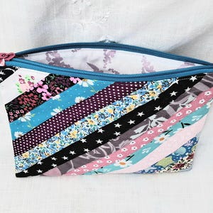 May include: A patchwork zippered pouch in a variety of patterned fabrics. The design features diagonal stripes in shades of blue, pink, black, and white, with floral, polka dot, and star prints. The zip is blue with a pink pull.